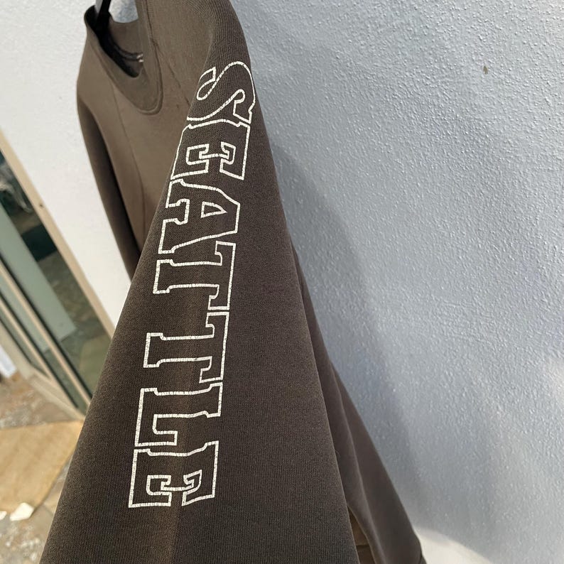 May include: A dark brown sweatshirt with the word "SEATTLE" printed vertically in white block letters on the left sleeve. The sweatshirt is hanging on a black hanger.