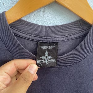 Op de afbeelding: Een donkergrijs T-shirt met een zwart label waarop "Local Motion Hawaiian Surfwear" staat en een maat L-label. Het label heeft ook de tekst "Care/Contents on Reverse Assembled in Jamaica of U.S.A. Parts".