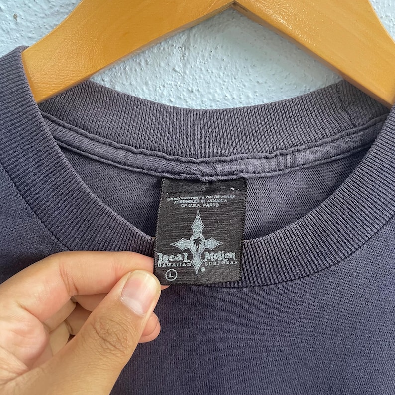Op de afbeelding: Een donkergrijs T-shirt met een zwart label waarop "Local Motion Hawaiian Surfwear" staat en een maat L-label. Het label heeft ook de tekst "Care/Contents on Reverse Assembled in Jamaica of U.S.A. Parts".
