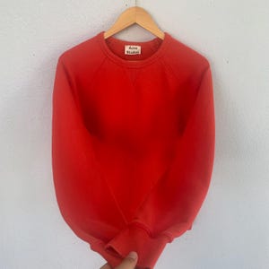 Op de afbeelding: Een felrood sweatshirt met een ronde hals en lange mouwen. Het sweatshirt hangt aan een houten hanger. Een klein label met de tekst "Acne Studios" is zichtbaar bij de halslijn.