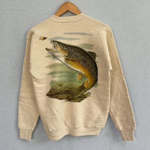 May include: A beige sweatshirt featuring a detailed illustration of a brown trout leaping upwards, with a fly above. The artwork includes shades of brown, yellow, and green, set against a light background. The sweatshirt is hanging on a wooden hanger.