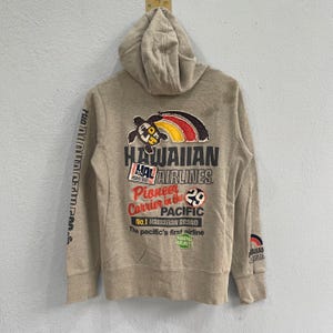 Puede incluir: Sudadera con capucha de color beige con capucha y bolsillo delantero. La parte trasera presenta un gráfico colorido con el texto "HAWAIIAN AIRLINES" y otro texto. La manga izquierda tiene texto vertical y la manga derecha un pequeño logotipo.