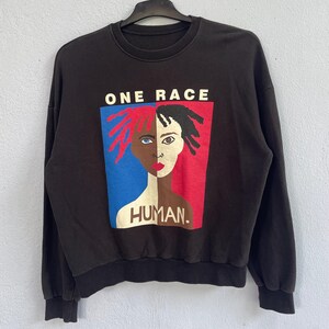 Op de afbeelding: Zwart sweatshirt met de tekst "ONE RACE HUMAN". De afbeelding toont een gespleten gezicht, de ene kant in blauw en rood, de andere kant in zwart en bruin. De sweatshirt heeft een ronde hals.