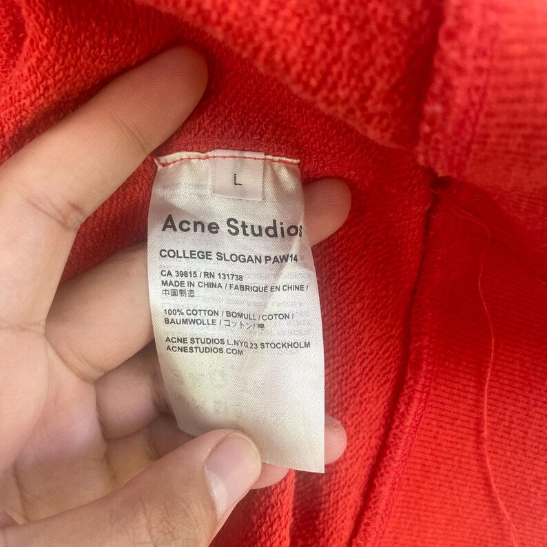 Op de afbeelding: Close-up van een rood kledingstuk met een wit label. Het label leest "Acne Studios COLLEGE SLOGAN PAW14" met details zoals "100% COTTON" en de website van het merk. Het kledingstuk is maat L.