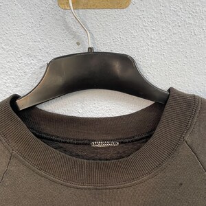 May include: A dark brown sweatshirt with a crew neck, suspended from a black hanger. The sweatshirt features a ribbed collar and cuffs. The material looks to be soft and comfortable.
