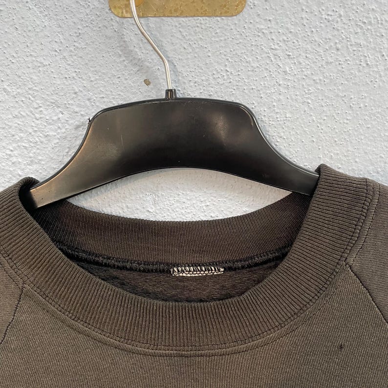 May include: A dark brown sweatshirt with a crew neck, suspended from a black hanger. The sweatshirt features a ribbed collar and cuffs. The material looks to be soft and comfortable.