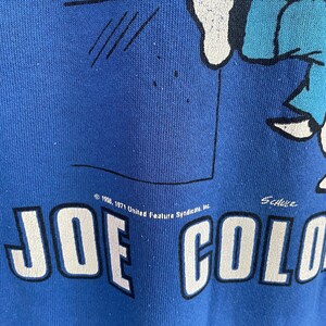 Peut inclure: Un sweat-shirt bleu avec une impression graphique d'un personnage de dessin anim&eacute;. Le texte indique JOE COOL en grandes lettres blanches avec des contours noirs. Au-dessus, on lit 1958, 1971 United Feature Syndicate, Inc. et la signature Schulz.