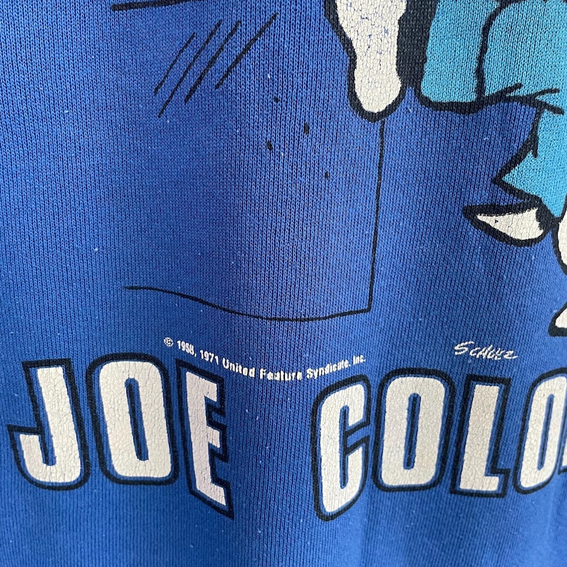 Peut inclure: Un sweat-shirt bleu avec une impression graphique d'un personnage de dessin anim&eacute;. Le texte indique JOE COOL en grandes lettres blanches avec des contours noirs. Au-dessus, on lit 1958, 1971 United Feature Syndicate, Inc. et la signature Schulz.