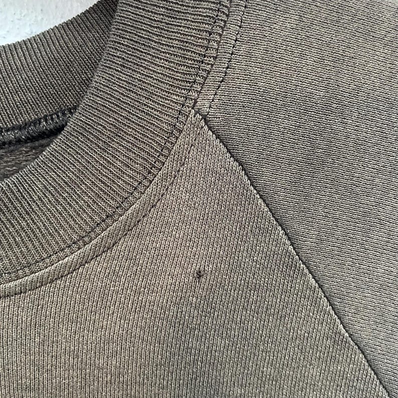 May include: A close-up shot of a dark olive green sweatshirt. The image focuses on the crew neck and shoulder seam, highlighting the fabric's texture and stitching. A small dark mark is visible on the fabric.