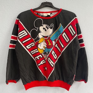 Puede incluir: Sudadera negra de manga larga con Mickey Mouse sosteniendo un balón de fútbol americano y una tabla de snowboard. El diseño incluye las palabras "Mickey Action" en un patrón diagonal rojo y blanco. La sudadera tiene detalles de rayas rojas y blancas en las mangas y un cuello y dobladillo rojos.