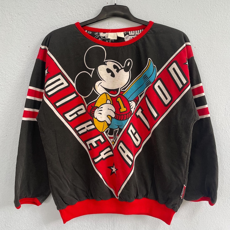 May include: Black long-sleeve sweatshirt featuring Mickey Mouse holding a football and a snowboard. The design includes the words "Mickey Action" in a red and white diagonal pattern. The sweatshirt has red and white striped accents on the sleeves and a red neckline and hem.