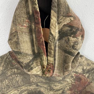 May include: Close-up of a camouflage hooded sweatshirt. The hood is made of a brown, green, and red camouflage pattern fabric. The hood has two metal snap closures at the neckline. The interior of the hood is black.