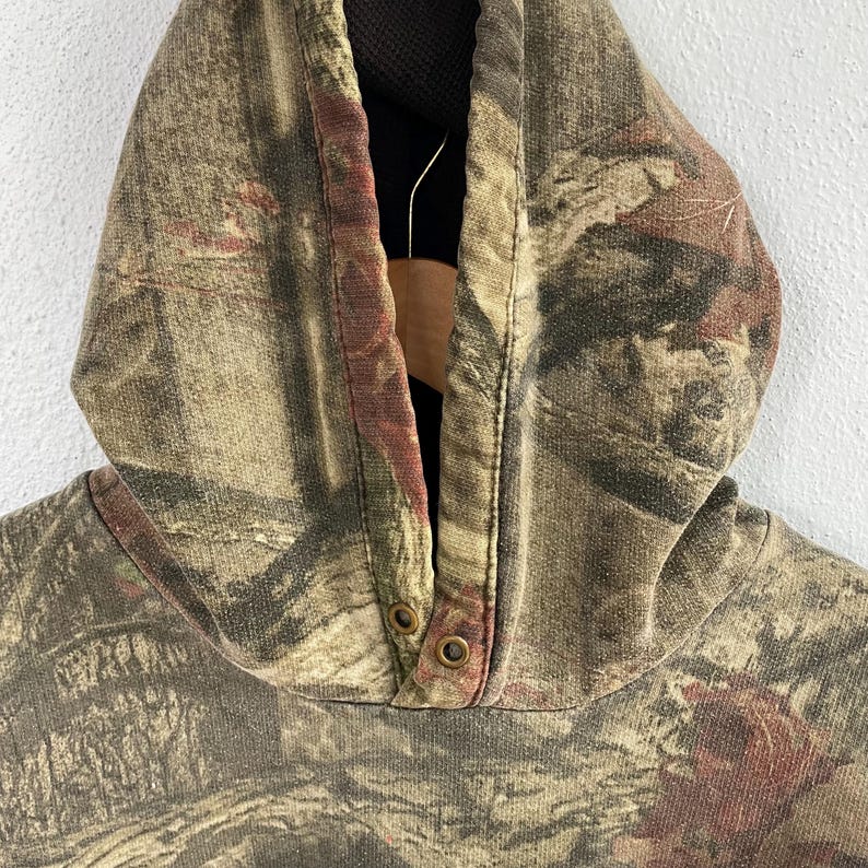 May include: Close-up of a camouflage hooded sweatshirt. The hood is made of a brown, green, and red camouflage pattern fabric. The hood has two metal snap closures at the neckline. The interior of the hood is black.