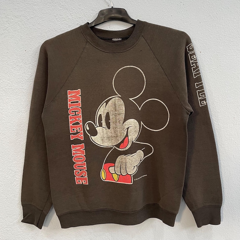 May include: A brown vintage sweatshirt with a distressed Mickey Mouse graphic. The sweatshirt features the words "Mickey Mouse" printed vertically on the left sleeve and "Special Tee" on the right sleeve. A classic Disney design.