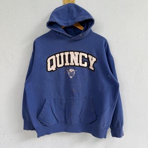 Może przedstawiać: Niebieska bluza z kapturem z napisem QUINCY dużymi białymi literami z czarnym obrysem na klatce piersiowej. Pod tekstem znajduje się mała grafika rogatego stworzenia. Odzież posiada kieszeń typu kangurka i widoczne są na niej plamy.