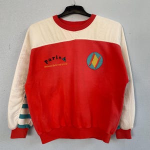 Può includere: Una felpa a maniche lunghe con corpo principale rosso e parte superiore del petto e maniche color crema. La parte anteriore mostra la scritta "paris" e "maison du le coq sportif" con una toppa circolare contenente un rombo giallo.