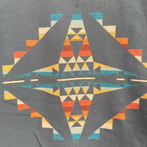 Może przedstawiać: Ciemnoszara koszulka z geometrycznym wzorem w odcieniach pomarańczu, ż&oacute;łci, niebieskiego i bieli. Symetryczny wz&oacute;r ma centralny kształt rombu i schodkowe krawędzie, co przywodzi na myśl styl południowo-zachodni lub plemienny.