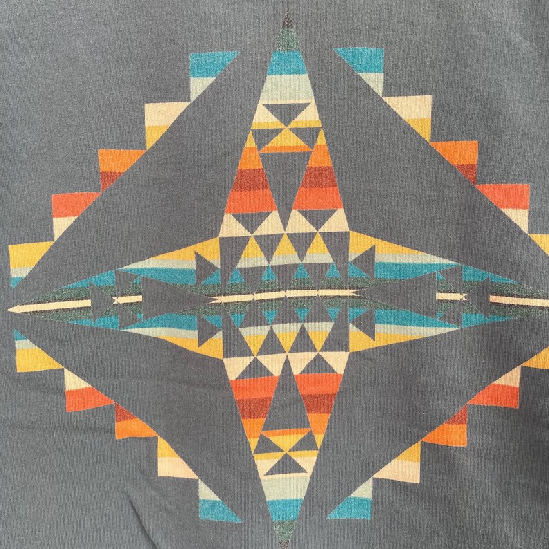 Może przedstawiać: Ciemnoszara koszulka z geometrycznym wzorem w odcieniach pomarańczu, ż&oacute;łci, niebieskiego i bieli. Symetryczny wz&oacute;r ma centralny kształt rombu i schodkowe krawędzie, co przywodzi na myśl styl południowo-zachodni lub plemienny.