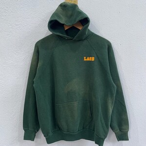 May include: A dark green hooded sweatshirt with the word "LASD" printed in orange on the chest. The sweatshirt has a front pocket and a drawstring hood. The fabric appears to be a worn, vintage style.