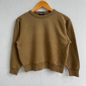 Pode incluir: Uma camisola castanha de gola redonda com punhos e bainha canelados, pendurada num cabide de madeira. O nome da marca "UNIQLO" é visível na gola. A camisola é feita de um material macio e de cor lisa.