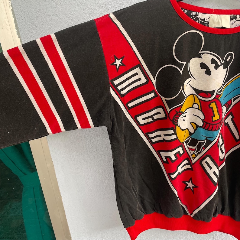 May include: Black vintage sweatshirt featuring Mickey Mouse holding a football. The sweatshirt has red and white stripes on the sleeves and the word "Mickey" in white lettering. The bottom and collar are trimmed in red.