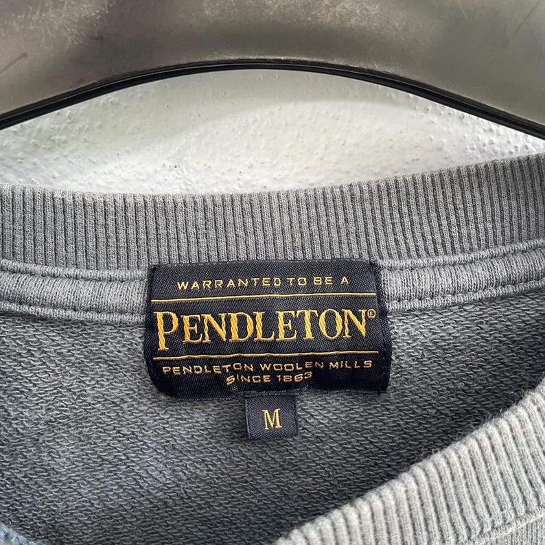 Może przedstawiać: Zbliżenie na szarą metkę bluzy Pendleton. Czarna metka głosi "Warranted to be a Pendleton" z napisem "Pendleton Woolen Mills Since 1863" poniżej. Rozmiar "M" jest wydrukowany poniżej. Bluza jest szara.