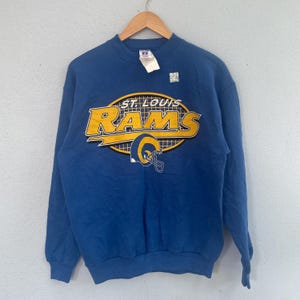 Pode incluir: Camisola azul com o texto "ST. LOUIS RAMS" em amarelo e branco. A camisola apresenta um gráfico de capacete de futebol americano amarelo. A peça de roupa está pendurada num cabide de madeira.