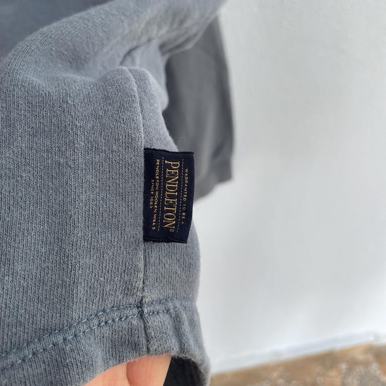 Może przedstawiać: Zbliżenie szarej koszulki z czarną etykietą. Na etykiecie widnieje napis "Pendleton" z tekstem "Warranted to be a Pendleton Woolen Mills Since 1863". Koszulka wykonana jest z miękkiego, wygodnego materiału.