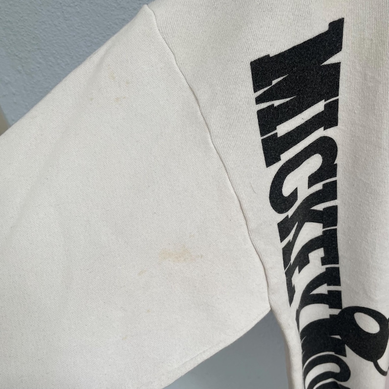Puede incluir: Una sudadera de manga larga color blanco hueso con la palabra MICKEY impresa verticalmente en letras may&uacute;sculas negras grandes en el lateral. La tela muestra manchas tenues de color marr&oacute;n claro en la manga cerca del hombro y la costura.