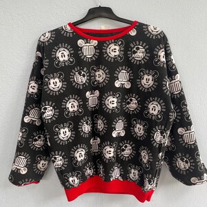 May include: Black sweatshirt with a repeating white Mickey Mouse pattern and red trim on the collar and hem. The design includes Mickey's face and other graphic elements. The long-sleeved top is hanging on a black hanger.