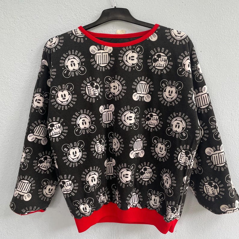 May include: Black sweatshirt with a repeating white Mickey Mouse pattern and red trim on the collar and hem. The design includes Mickey's face and other graphic elements. The long-sleeved top is hanging on a black hanger.