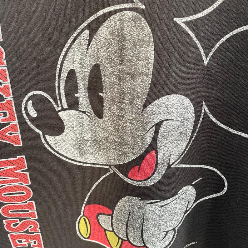 May include: Close-up of a black t-shirt featuring a distressed graphic of Mickey Mouse. The iconic character's face is in silver with red and yellow accents. The words "Mickey Mouse" are printed in red.