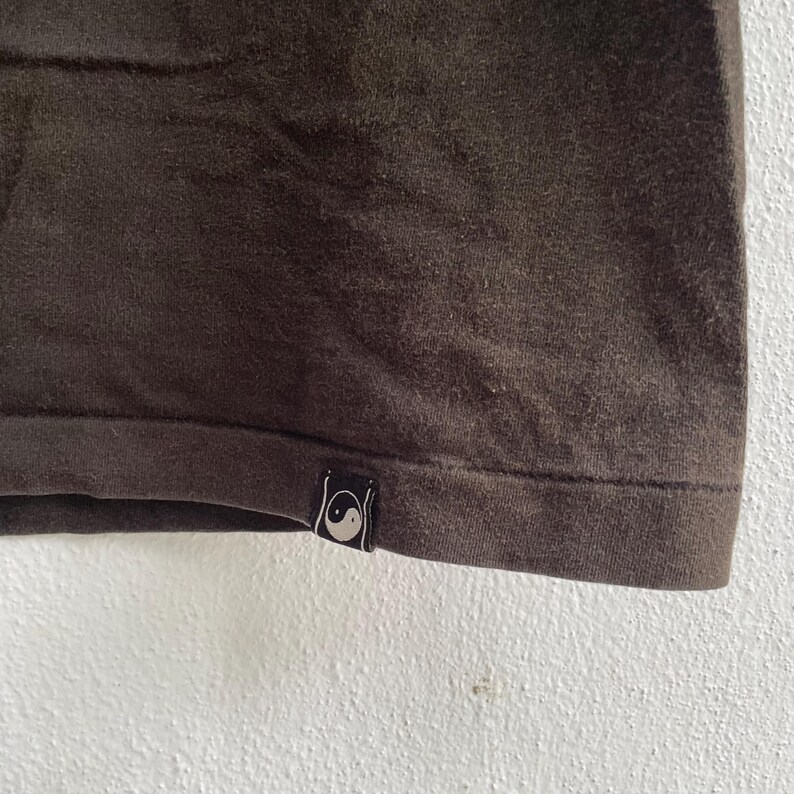 Puede incluir: Primer plano de una camiseta marr&oacute;n oscuro. El dobladillo inferior es visible, con una peque&ntilde;a etiqueta negra con un s&iacute;mbolo de yin-yang blanco. La tela tiene un aspecto ligeramente texturizado.