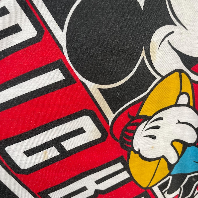 May include: Close-up of a white t-shirt featuring a large graphic of Mickey Mouse. The design includes the word "GO" in white block letters on a red background. Mickey Mouse is holding a yellow football.