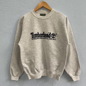 May include: A light gray Timberland sweatshirt with a crew neck and long sleeves. The black Timberland logo and text "WIND.WATER.EARTH.AND SKY" are printed on the front. The sweatshirt is hanging on a wooden hanger.