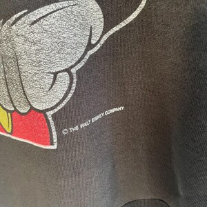May include: Close-up of a black sweatshirt with a Mickey Mouse graphic. The design shows a white hand with red and yellow accents. The text "THE WALT DISNEY COMPANY" is printed in white.