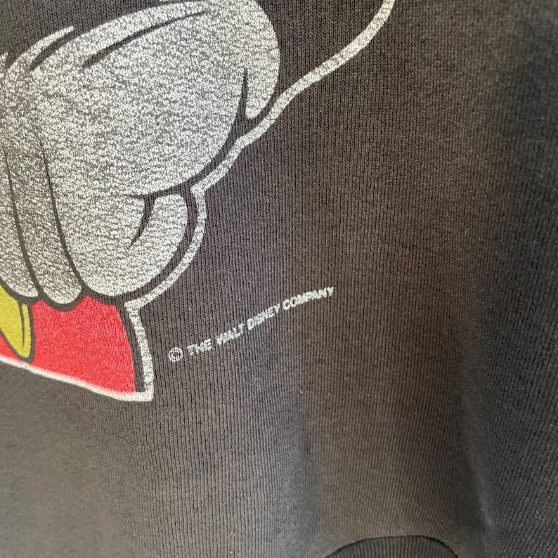 May include: Close-up of a black sweatshirt with a Mickey Mouse graphic. The design shows a white hand with red and yellow accents. The text "THE WALT DISNEY COMPANY" is printed in white.