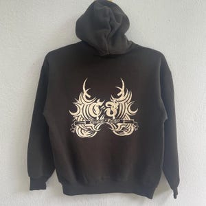 May include: A dark brown hooded sweatshirt with a cream-coloured graphic on the back. The graphic features a stylized design with the text "Christian Fletcher" on a banner. The sweatshirt has a hood and long sleeves.