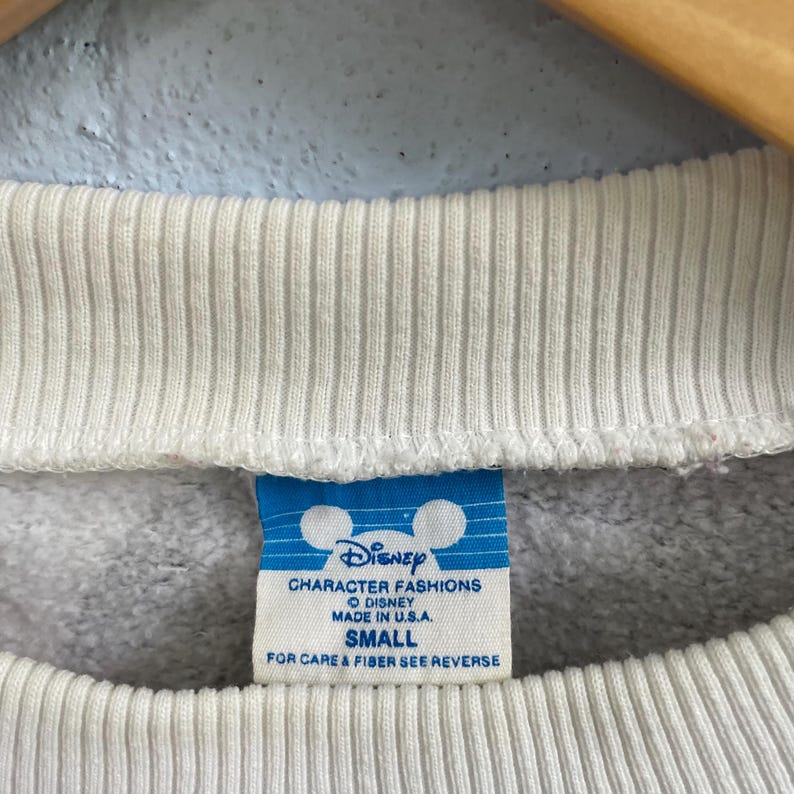 Puede incluir: Un cuello acanalado blanco en una sudadera gris claro con una etiqueta azul y blanca que presenta el logotipo de Disney, el texto Character Fashions, Made in U.S.A. y la talla Small. La etiqueta tambi&eacute;n incluye instrucciones de cuidado.