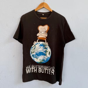 May include: A black short-sleeved t-shirt featuring a graphic of a cartoon slice of bread standing on planet Earth. Below the graphic, the text reads WE BUTTER THE BREAD WITH BUTTER in white capital letters. The shirt has a crew neckline.