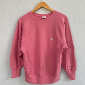 May include: A pink Champion sweatshirt with a crew neck and long sleeves. The sweatshirt features the Champion logo on the chest and left sleeve. The garment is hanging on a wooden hanger against a light-colored wall.