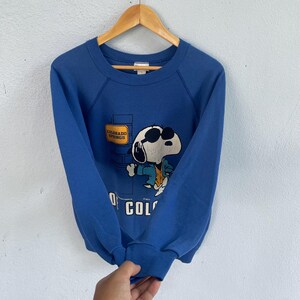 Peut inclure: Un sweat-shirt bleu &agrave; manches longues avec un motif repr&eacute;sentant un chien de dessin anim&eacute; portant des lunettes de soleil et une veste. Le motif inclut un panneau jaune indiquant Colorado Springs et le texte partiel Colorado en bas.
