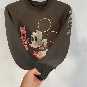 May include: A dark brown sweatshirt with a distressed Mickey Mouse design on the front. The sleeves display the text "SPECIAL ISSUE" and "MICKEY MOUSE" in red and white. It has a crew neck and ribbed cuffs.