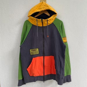 Puede incluir: Una sudadera con capucha con cremallera Fubu multicolor. La sudadera con capucha presenta una capucha amarilla, mangas verdes, un cuerpo azul marino y un bolsillo naranja. El logotipo de Fubu es visible en la parte delantera. La sudadera tiene cremallera completa y cordones.