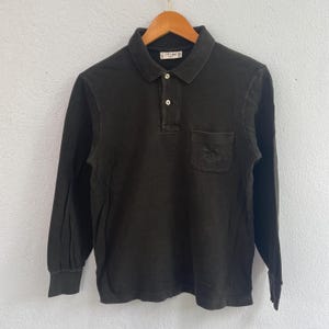Puede incluir: Un polo de manga larga verde oliva oscuro con cuello y bolsillo en el pecho izquierdo. La camisa tiene una tapeta de tres botones y un pequeño logotipo cerca del cuello. La camisa está colgada en una percha de madera.