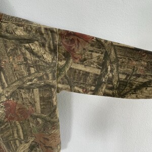 May include: Close-up of a long-sleeved shirt with a camouflage pattern. The design features a blend of brown, green, and red hues, mimicking a forest environment. The sleeve is visible, showcasing the fabric's texture and the stitching along the seam.