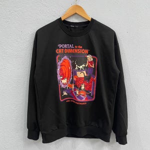 Puede incluir: Sudadera negra con un gráfico colorido que dice "PORTAL to the CAT DIMENSION" con una ilustración de dibujos animados de gatos, un portal y una persona. La frase "AND OTHER NIGHTMARES!" también está impresa en la sudadera.