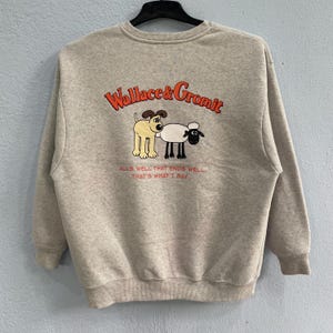 May include: A light grey sweatshirt with the Wallace & Gromit logo in orange. The design features a cartoon dog and sheep. Below the characters is the phrase "ALL'S WELL THAT ENDS WELL, THAT'S WHAT I SAY." The sweatshirt is a casual, comfortable garment.