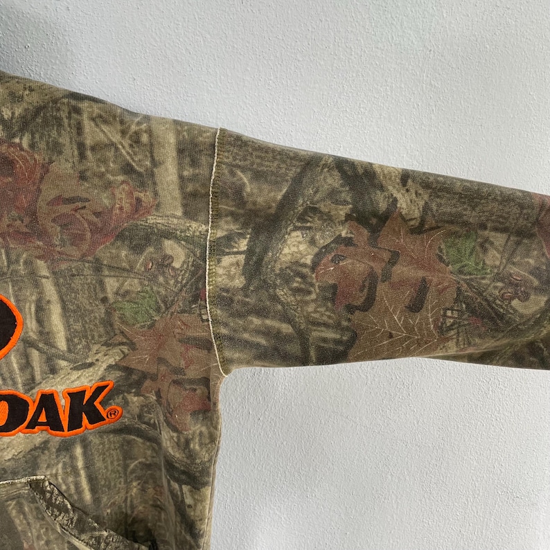 May include: A camouflage-patterned hoodie with the word "DAK" in orange block letters. The hoodie features a drawstring hood and a kangaroo pocket. The fabric has a brown, green, and tan color scheme, with a leaf and tree branch design.