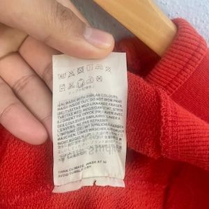 Op de afbeelding: Close-up van een rood kledingstuk met een wit etiket. Het etiket bevat wasvoorschriften in meerdere talen, met was- en droogsymbolen. De tekst bevat zinnen als "Wassen met dezelfde kleuren" en "Niet in de droger."